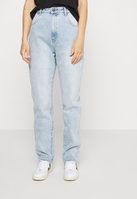 Jeans de cós alto azul claro com bolsos laterais, fecho de botão e um corte solto, combinados com ténis brancos com detalhes em preto.