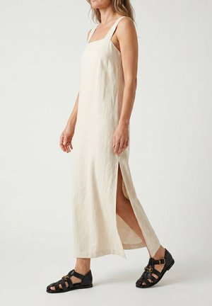 Vrouw draagt een mouwloze beige linnen maxi-jurk met een zijsplit en zwarte leren platte sandalen met gespdetail.