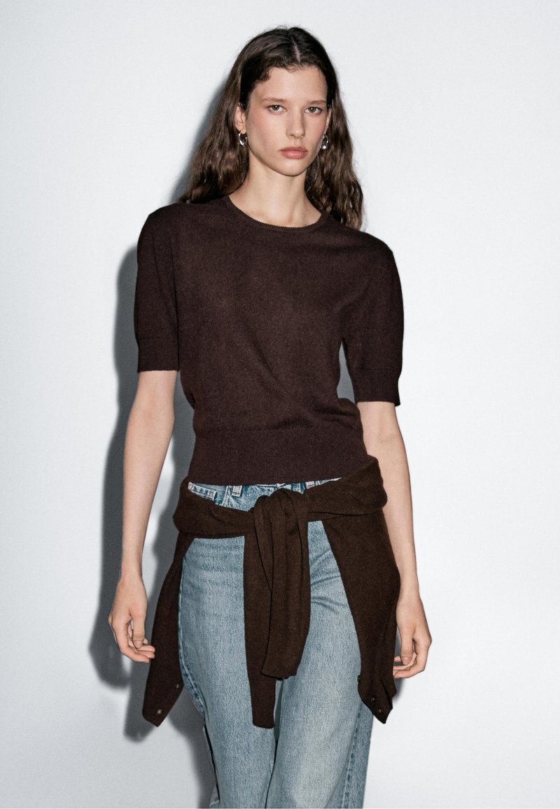 Bruine short-sleeve trui gemaakt van zacht materiaal, met een ronde hals. Gekleed met lichtblauwe jeans en een gebonden bruine trui om de taille.