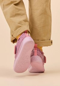 Chaussures pour enfants roses avec une texture pelucheuse, une sangle Velcro et des semelles en caoutchouc, associées à un pantalon beige et une chaussette jaune, sur un fond neutre.