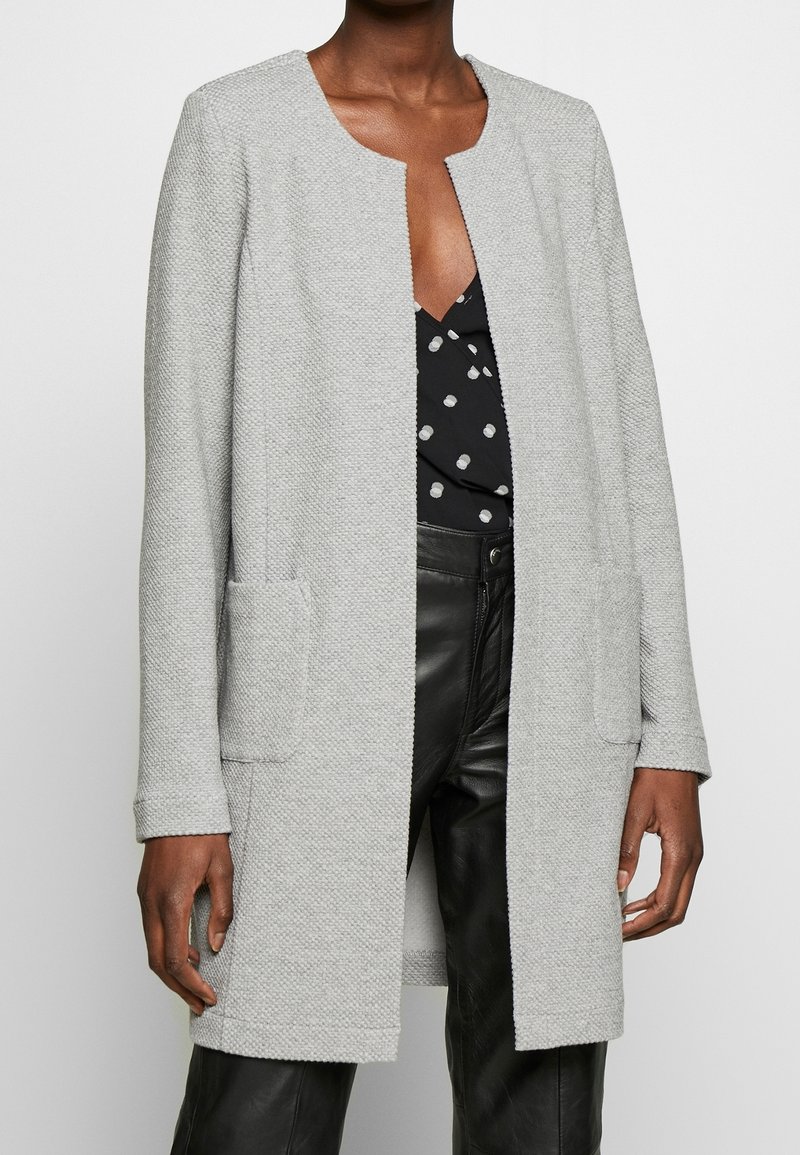 Cappotto grigio chiaro intrecciato con scollatura rotonda, apertura frontale, due tasche laterali e maniche lunghe. Indossato sopra una maglietta nera a pois e pantaloni in pelle.