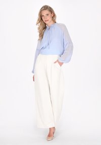 Helles blaues Blusenshirt mit gepunktetem Muster und durchsichtigen Puffärmeln, kombiniert mit weißen, weit geschnittenen Hosen. Das Model hat eine Hand in der Tasche.