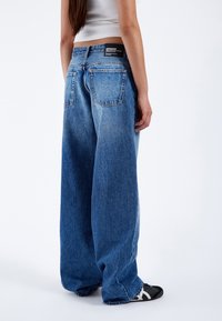 Wijd-uitlopende blauwe denim jeans met een vervaagde wassing, vijf zakken en een zwart merkje op de achterste tailleband. Gladde textuur, casual ontwerp.