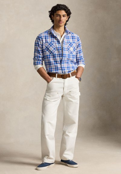 Muškarac obučen u plavu kariranu košulju, bijele hlače s smeđim remenom, plave tenisice, stoji s rukama u džepovima ispred neutralne pozadine.
