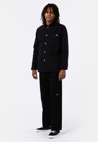 Veste en velours côtelé noire avec un devant à boutons, deux poches avant et un petit logo sur la poitrine. Associée avec un pantalon noir et des baskets noires.