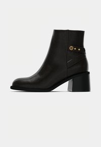 Botas de tornozelo castanhas com um acabamento texturizado, apresentando um cano curvado, detalhes em dourado e um salto bloco para maior estabilidade.