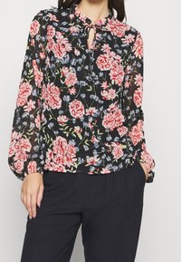Blusa floreale con maniche lunghe e fluenti, sfondo nero e fiori rosso-rosa. Presenta un collo rotondo con un'apertura a chiave.