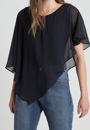 Blouse noire asymétrique en voile avec manches kimono courtes et fluides, portée avec un jean bleu taille haute sur une personne debout.