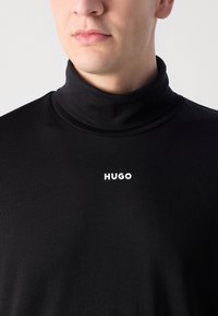 Svart turtleneck genser med en ribbestrikket tekstur og hvit "HUGO" branding på brystet. Har en tettsittende design og høy krage.