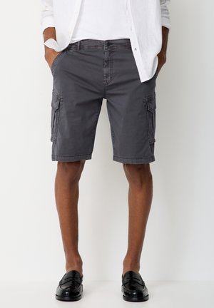 Man wearing dark gray cargo shorts, white shirt, and black loafers, standing with hands in pockets against a plain background.