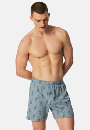 Uomo senza maglietta con capelli corti che indossa pantaloncini grigi decorati con piccoli ananas, in piedi con le mani dietro la schiena e lo sguardo rivolto verso il basso.