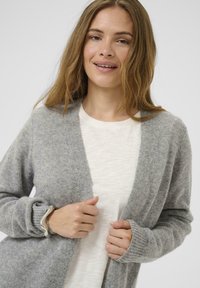 Grauer gestrickter Cardigan mit offener Vorderseite, gerippten Bündchen und strukturierter Oberfläche, getragen über einem hellen Oberteil. Hat eine lockere Passform.