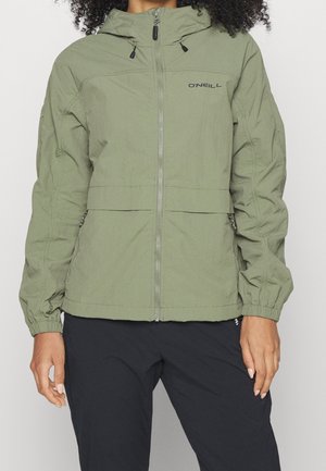 Femme portant une veste zippée O'Neill vert clair avec des poignets élastiques et un pantalon noir, montrée du cou aux hanches sur un fond uni.