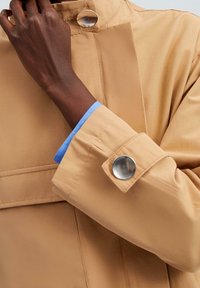Beżowa kurtka z wysokim kołnierzem, błyszczącym srebrnym guzikiem i niebieskim wykończeniem rękawów. Gładka faktura i minimalistyczny design.