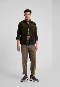 Καφέ μπουφάν από κορντόρι με δύο τσέπες στο στήθος, πλεκτό πουλόβερ με σχέδια, μπεζ παντελόνι με στενή γραμμή, και καφέ/μπλε αθλητικά παπούτσια με λευκές σόλες.