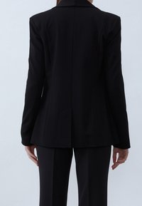 Czarna, dopasowana marynarka z pojedynczym rozcięciem z tyłu, gładka tkanina, klapy z wycięciami oraz usztywnione ramiona. Stylizowana z dopasowanymi spodniami.