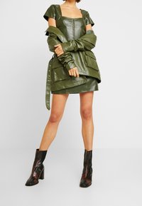 Grünes Kunstlederkleid mit quadratischem Ausschnitt, kurzen Ärmeln und geschichteter Gestaltung. Kombiniert mit schwarz-braunen gemusterten Stiefeletten.