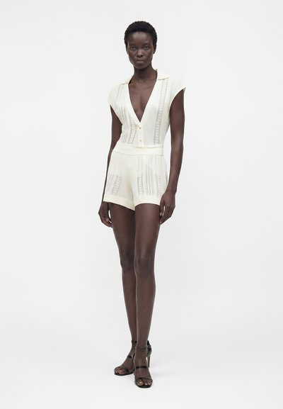 Γυναίκα που φοράει ένα εκρού ολόσωμο πλεκτό κοντομάνικο φόρεμα με κάθετες δαντελένιες λεπτομέρειες και μαύρα σανδάλια με ψηλό τακούνι, σε λευκό φόντο.