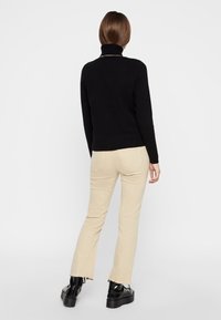 Schwarzer Rollkragenpullover mit taillierter Silhouette, kombiniert mit beigen, ausgestellten Hosen mit rohem Saum und schwarzen, klobigen Stiefeln mit Absatz.