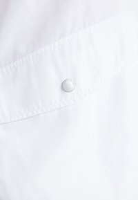 Biała tkanina o gładkiej fakturze, z klapą z srebrnym napem z wygrawerowanym napisem "TOMMY JEANS". Brak dodatkowych wzorów lub akcentów.