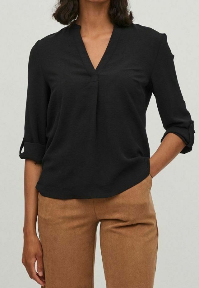 Femme portant un chemisier noir à col en V avec manches retroussées et pantalon couleur camel, main dans la poche, sur un fond neutre.