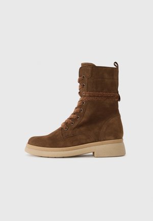 Bottine à lacets en daim marron avec semelle en caoutchouc beige clair et détail en laine autour de la cheville et des lacets, vue de côté sur fond uni.