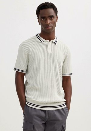 Joven con una camisa polo blanca acanalada con cuello y mangas con rayas negras, y pantalones cargo grises, de pie con las manos en los bolsillos.