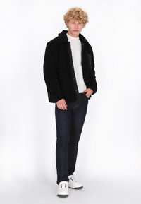 Giovane con capelli biondi ricci che indossa una giacca nera a trama, camicia bianca, jeans scuri e sneakers bianche, in piedi con una mano in tasca.