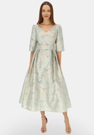 Kobieta ubrana w jasnoniebiesko-beżową sukienkę midi z kwiatowym wzorem dżakardowym, z rękawami do łokcia i dekoltem w serek, w połączeniu z cielistymi szpilkami na kostkę.