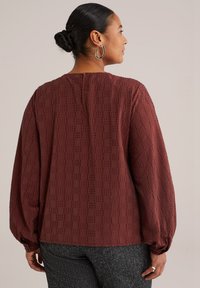 Blusa texturizada en color burdeos con mangas largas abullonadas, escote redondo y abertura en la parte posterior. Presenta un pequeño patrón geométrico y puños abotonados.
