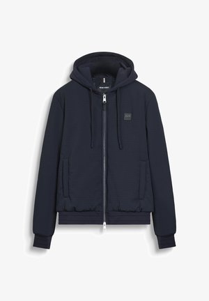 Felpa con zip trapuntata blu navy, dotata di lacci, tasche frontali, polsini e orlo a coste, e una piccola toppa con logo sul petto.