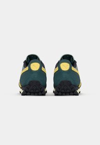 Pari teal ja keltainen Puma-tenniskenkiä nähtynä takaa mustilla piikkipohjilla valkoisella taustalla.