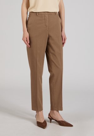 Kobieta w beżowym topie bez rękawów, brązowych prostych spodniach i brązowych niskich butach na obcasie z zaostrzonym noskiem na neutralnym tle.