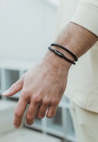 Brățări împletite negre pe o încheietură, cu un accent cilindric neted. Materialul pare a fi piele sau un textil similar.