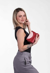 Red leather wallet with a textured pink accent and a fold-over design. Model holds it in a pose, wearing a black top and grey trousers.