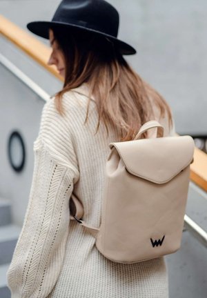 Beige leather backpack with a flap closure, rectangular shape, and a logo detail at the bottom. Worn over a cream knit sweater.