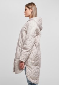 Polstret hettejakke i lys beige med diagonalt quiltet mønster, sidelommer og buet nederkant. Myk stoff med en glatt tekstur.