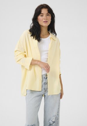 Yellow button-up shirt with long sleeves, made of soft fabric, paired with a white tank top and light blue distressed jeans.