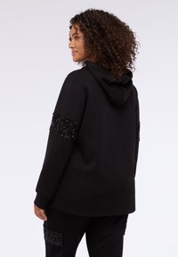 Felpa nera con cappuccio, caratterizzata da applicazioni di paillettes nere testurizzate sulle maniche. Materiale morbido, vestibilità comoda.