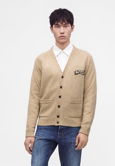 Jeune homme portant un cardigan beige boutonné sur une chemise blanche et un jean bleu, debout devant un fond blanc uni.