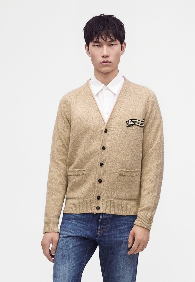 Jeune homme portant un cardigan beige boutonné sur une chemise blanche et un jean bleu, debout devant un fond blanc uni.