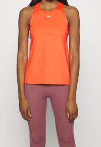 Heldere oranje mouwloze sporttop met een ronde hals en subtiele logo-details; gecombineerd met strakke paarse leggings met naadaccenten.