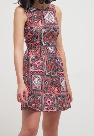 Woman wearing sleeveless red and black patterned dress with geometric and floral square designs, standing against a plain light background.