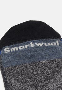 Sive in modro vzorčasta nogavica s črnim rebrastim robom, z belim logotipom "Smartwool" na vrhu. Mehak, pleten material.