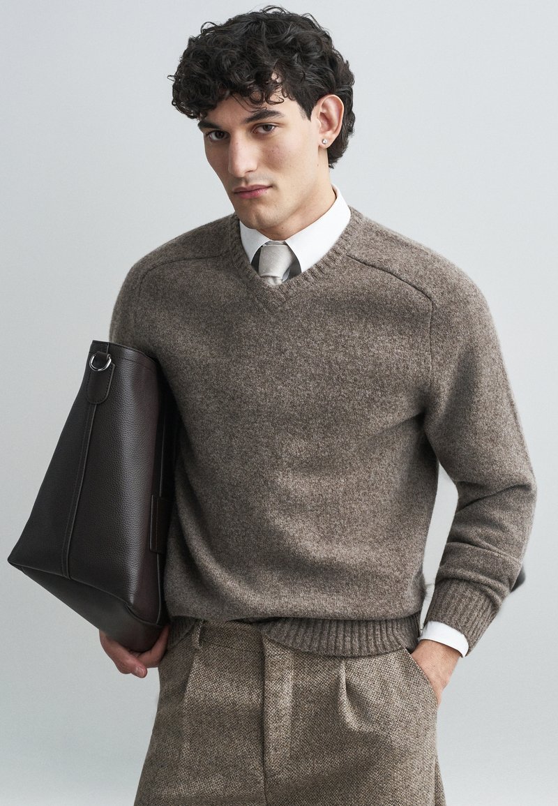 Maglione a V in lana marrone con orlo a costine, abbinato a una camicia grigia chiaro e cravatta. Tiene in mano una borsa in pelle marrone scuro con texture.