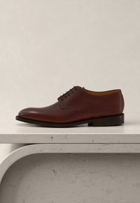 Καφέ δερμάτινο ανδρικό επίσημο παπούτσι με κορδόνια, τοποθετημένο πάνω σε μπεζ υφή λίθινης επιφάνειας, με φόντο ανοιχτό μπεζ χρώμα.