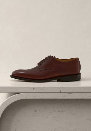 Zapato de vestir de hombre de cuero marrón con cordones, descansando sobre una superficie de piedra texturizada beige contra un fondo beige claro.