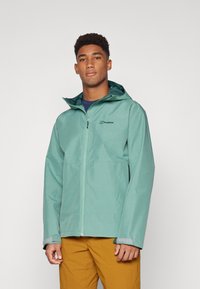 Chaqueta impermeable de color verde claro, fabricada en material sintético, con cremallera frontal, capucha ajustable y puños elásticos. Presenta un logo en el pecho.