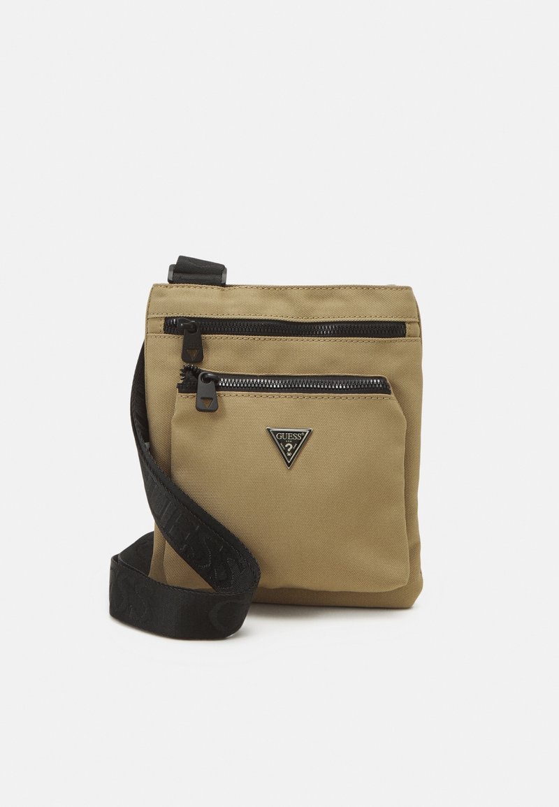 Sac bandoulière beige avec deux compartiments zippés, bandoulière noire ajustable avec logo embossé, et logo triangulaire Guess sur la poche avant.