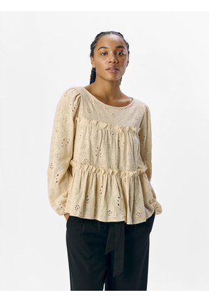 Blusa beige a maniche lunghe con scollatura rotonda, design a strati con balze, motivo a fiori con occhietti, e polsini arricciati. Abbinata a pantaloni neri.